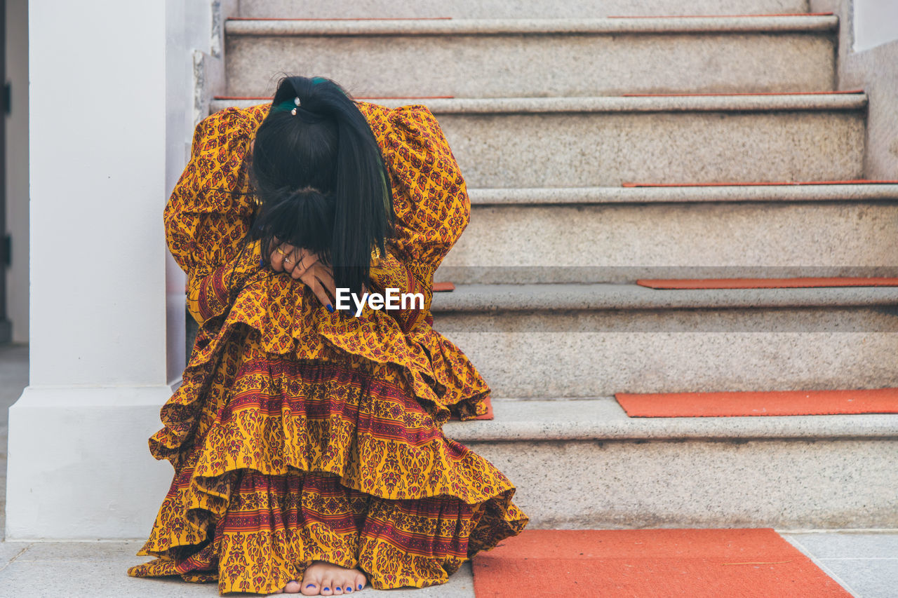 Asian woman wearing a yellow thai patterned dress sitting, she hide her face with her hands 
