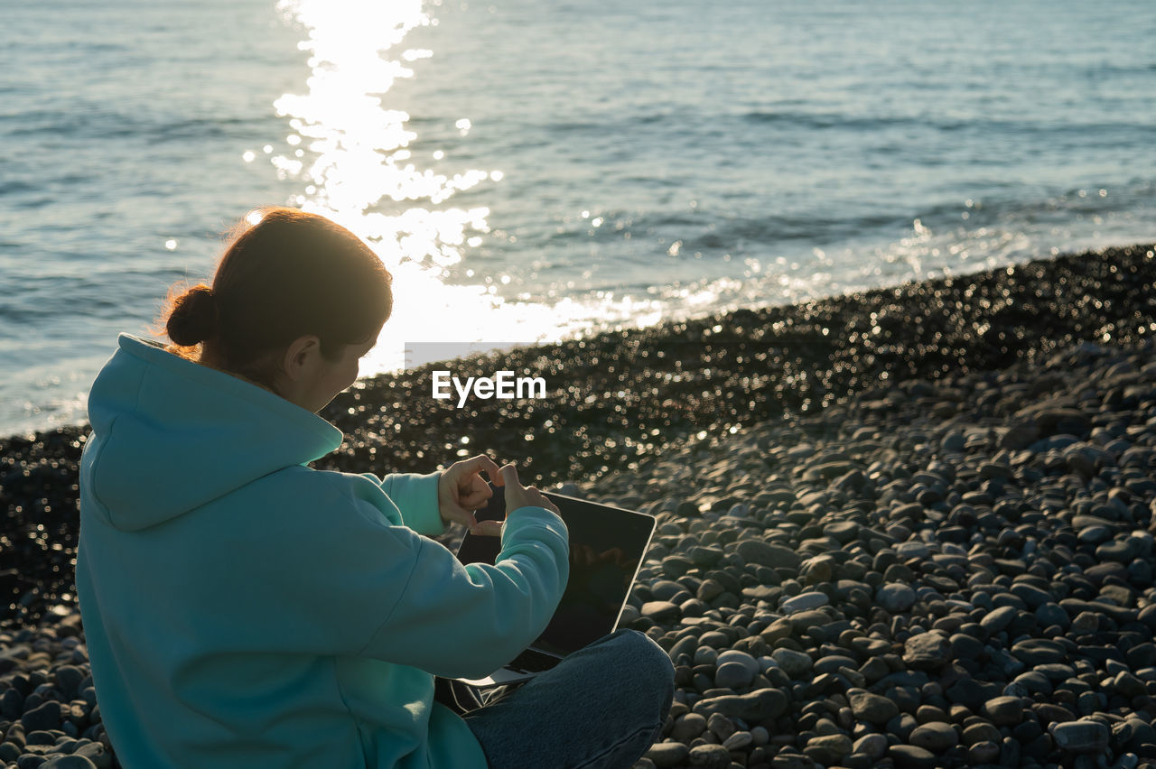 Woman sitting with laptop by sea on sunny day