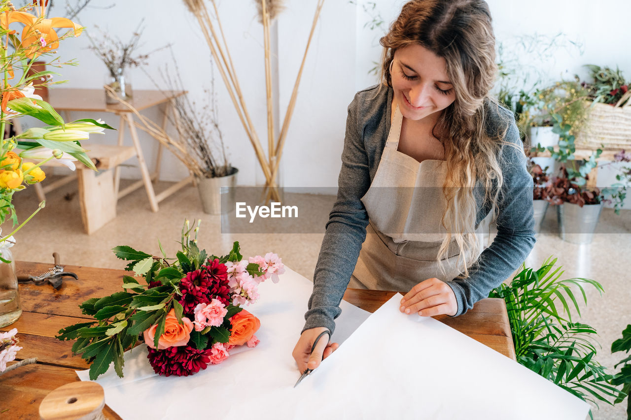 From above of crop happy ethnic young female florist in apron smiling and cutting wrapping paper while arranging elegant bouquet of fresh assorted colorful flowers