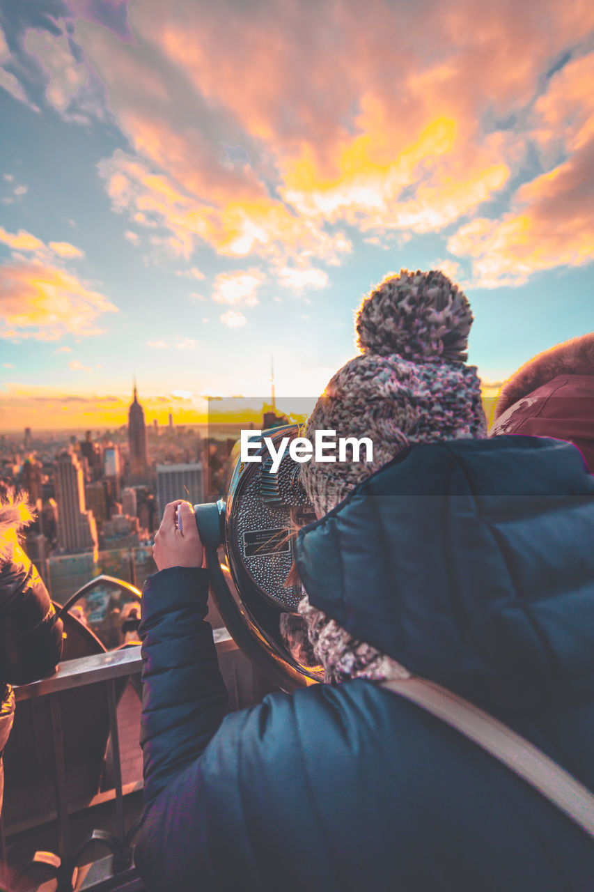 Woman looking through coin-operated binoculars against sky during sunset