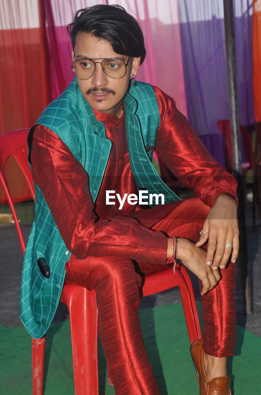 A indian young guy sitting on chair with wearing kurta, pajama, nehru coat with looking sideways 