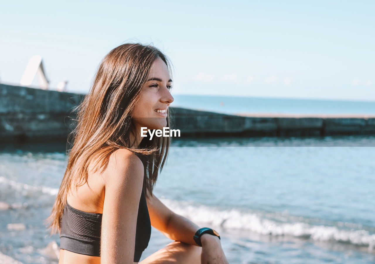 Woman looking away while sitting at beach against sky