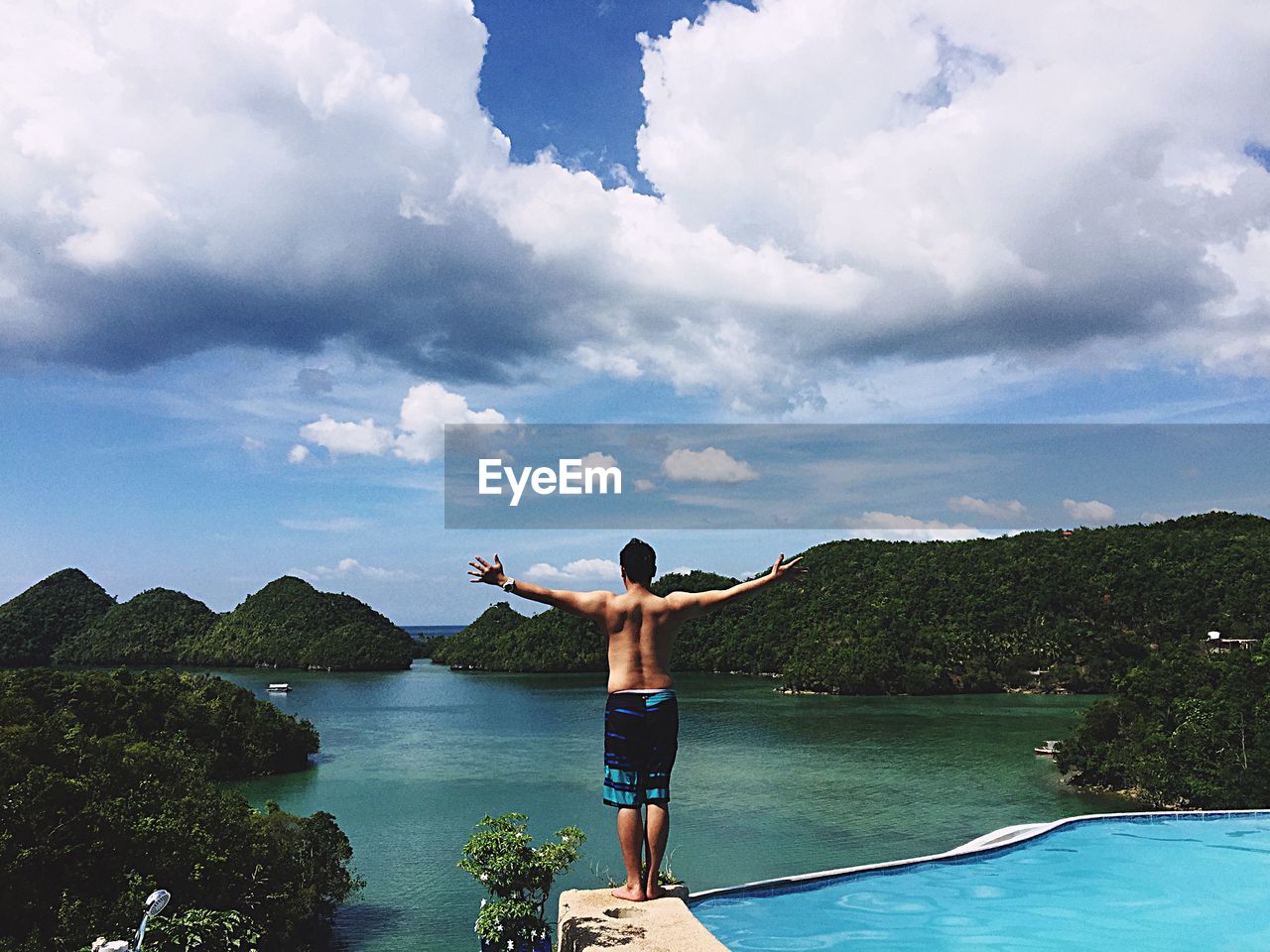 Rear view of shirtless with arms outstretched standing by infinity pool and sea against cloudy sky