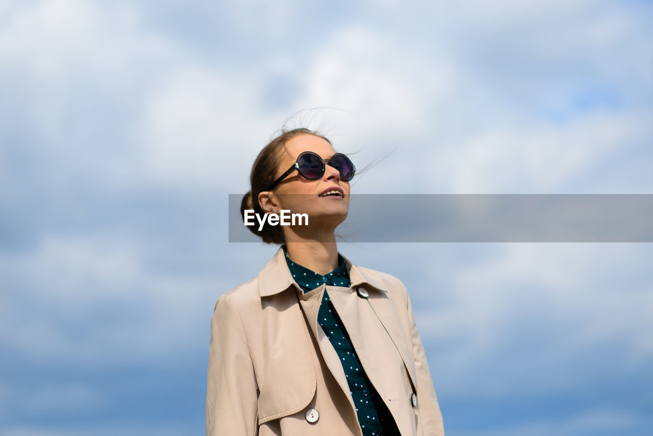 PORTRAIT OF YOUNG WOMAN STANDING AGAINST SKY