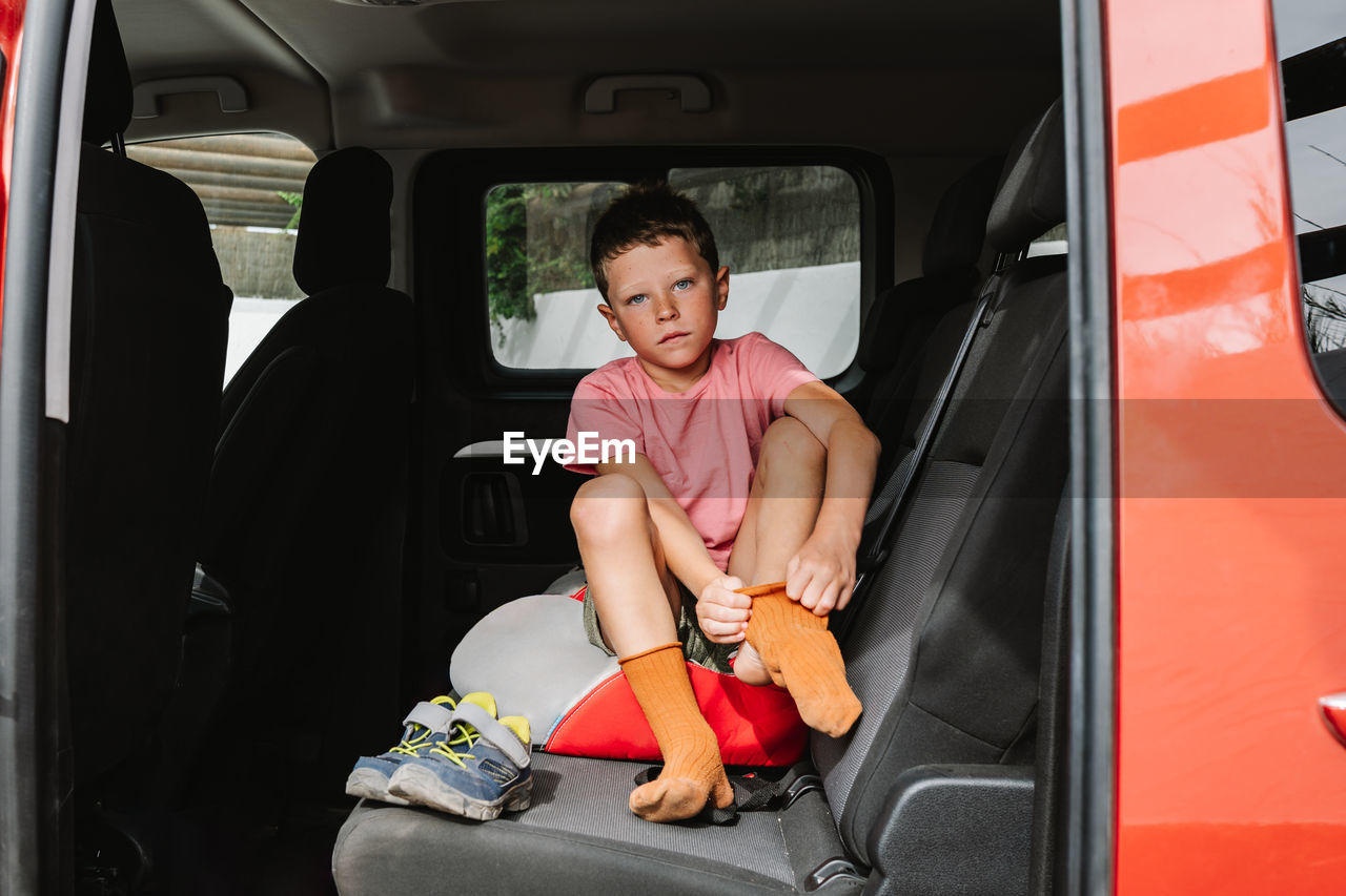 Full body kid in pink t shirt putting on orange socks and looking at camera while sitting on back seat of car during road trip