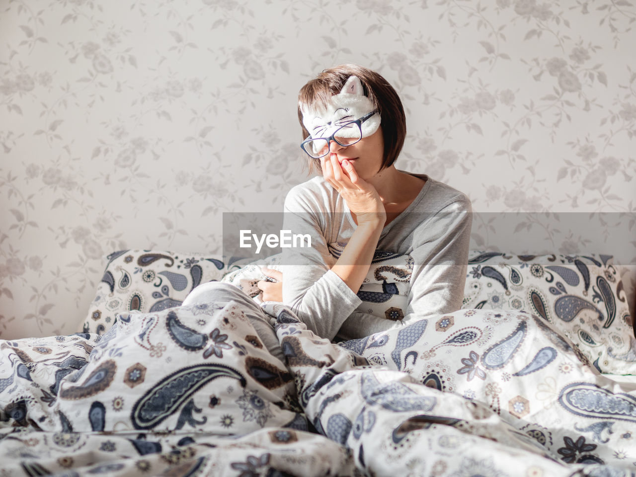 Young woman in grey pajama, glasses and sleeping mask in shape of cute sleeping cat face. 