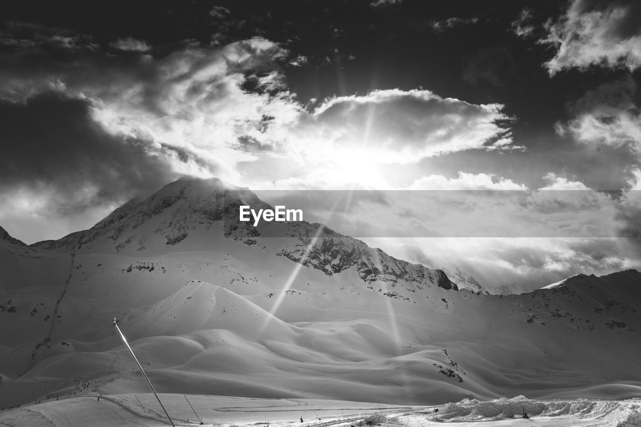 Black and white of the sun shining through the clouds above a ski slope in les arcs