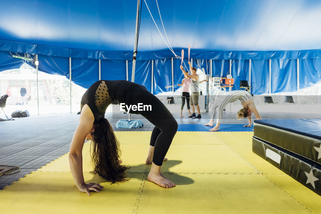Female artist bending over backwards while practicing with athletes in background at circus