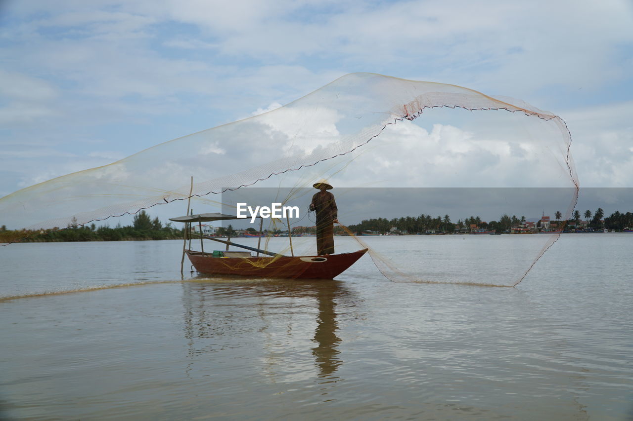 Fisherman throwing net while standing in boat on sea against sky