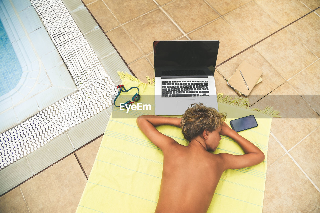 High angle view of boy sleeping by laptop and phone on beach towel at poolside