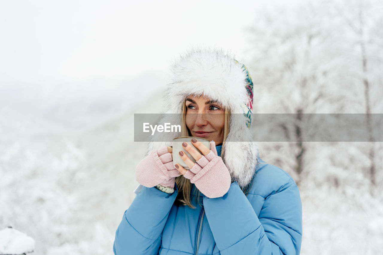 Portrait of a beautiful girl in a hat with earflaps and mittens outside holding a mug with a drink