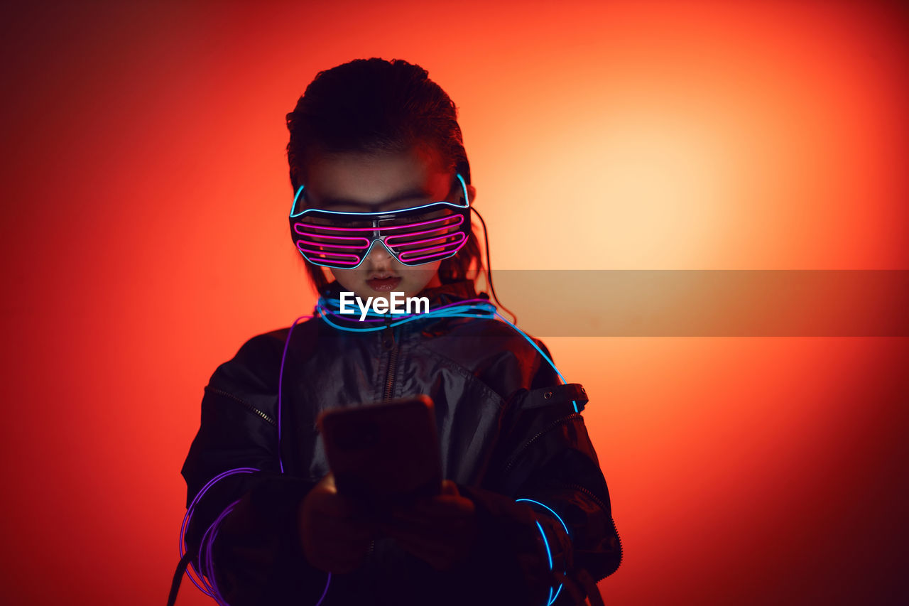 Portrait of robot boy child from the future in neon glasses and jacket looking at the phone at night