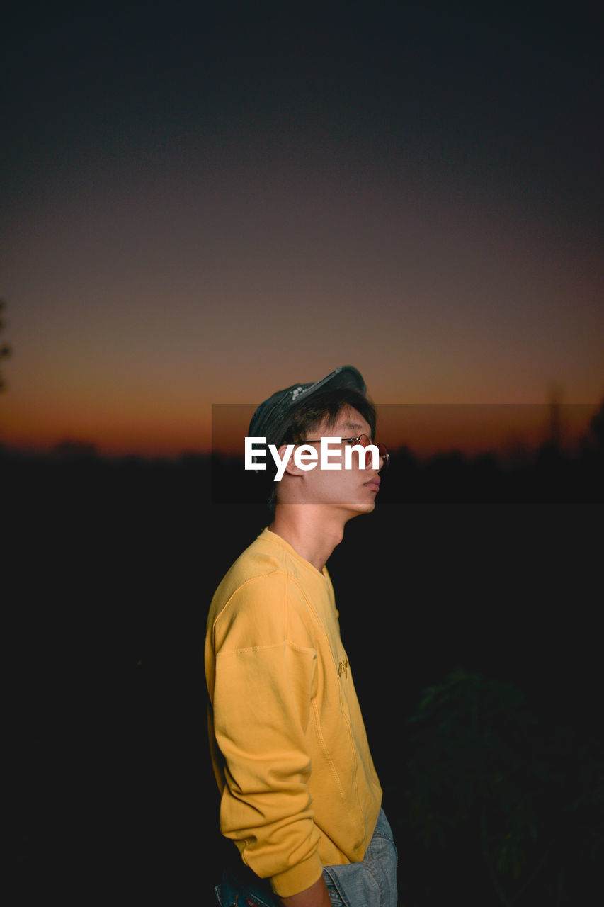 Side view of young man wearing sunglasses standing against sky at dusk