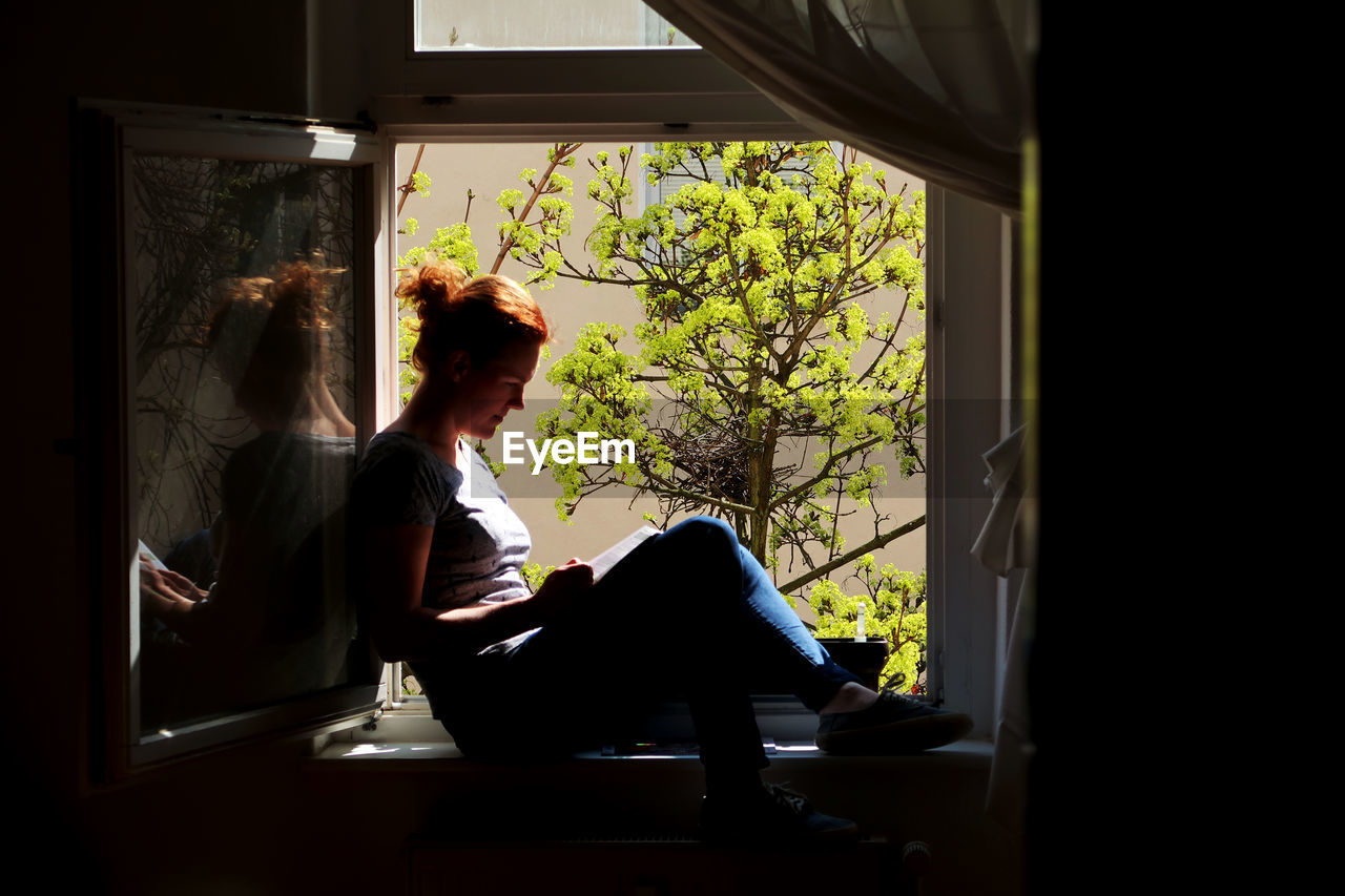 Side view of mid adult woman reading book while sitting on window sill at home