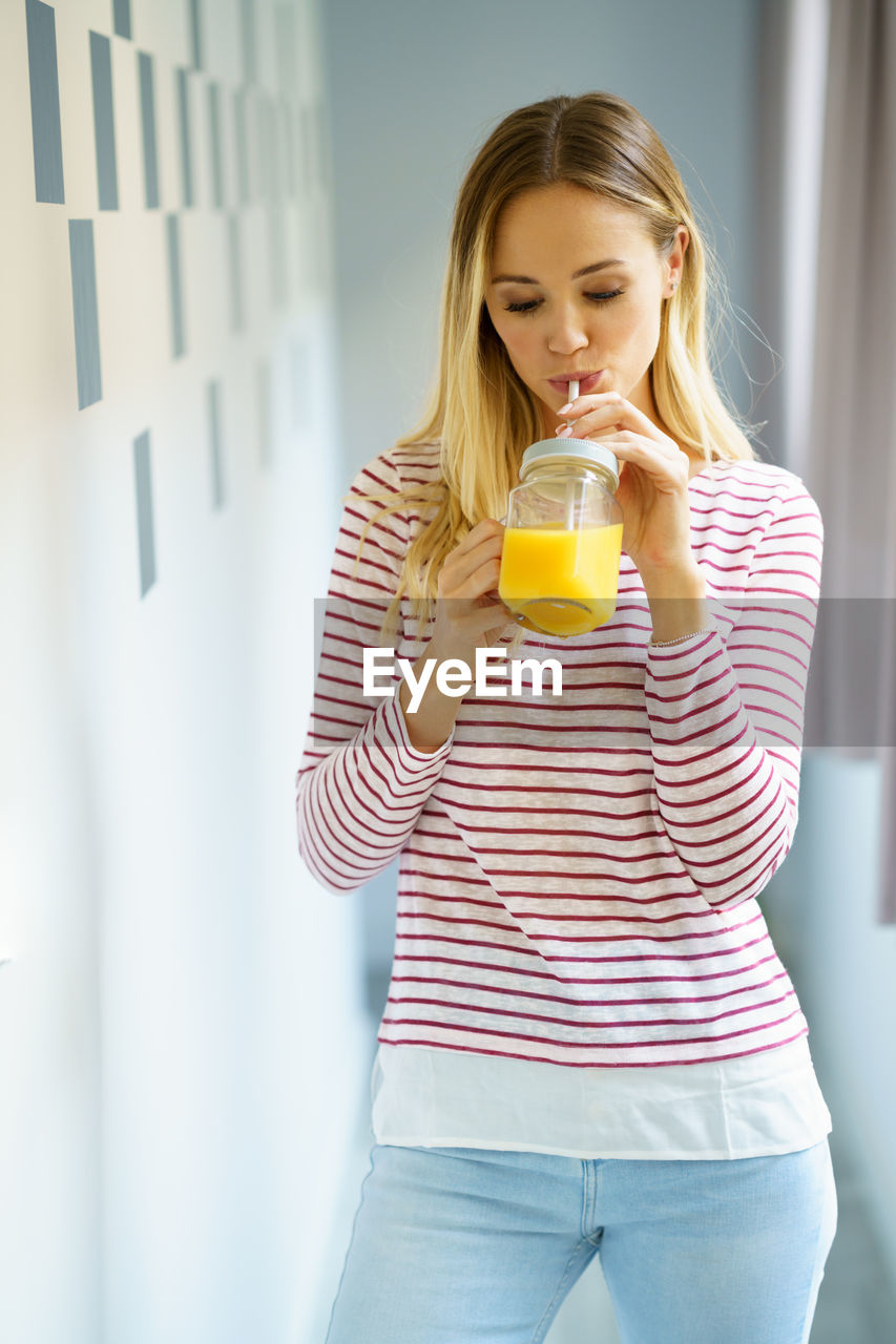 Young woman drinking juice while standing against wall