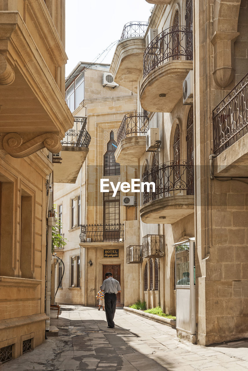 REAR VIEW OF MAN WALKING ON ALLEY AMIDST BUILDINGS