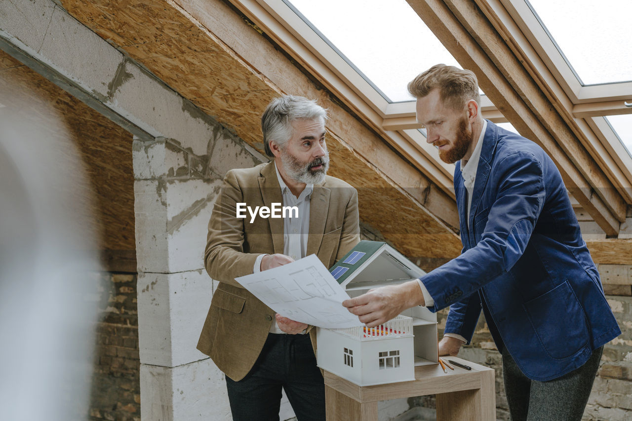 Architect explaining colleague over blueprint standing by house model at construction site