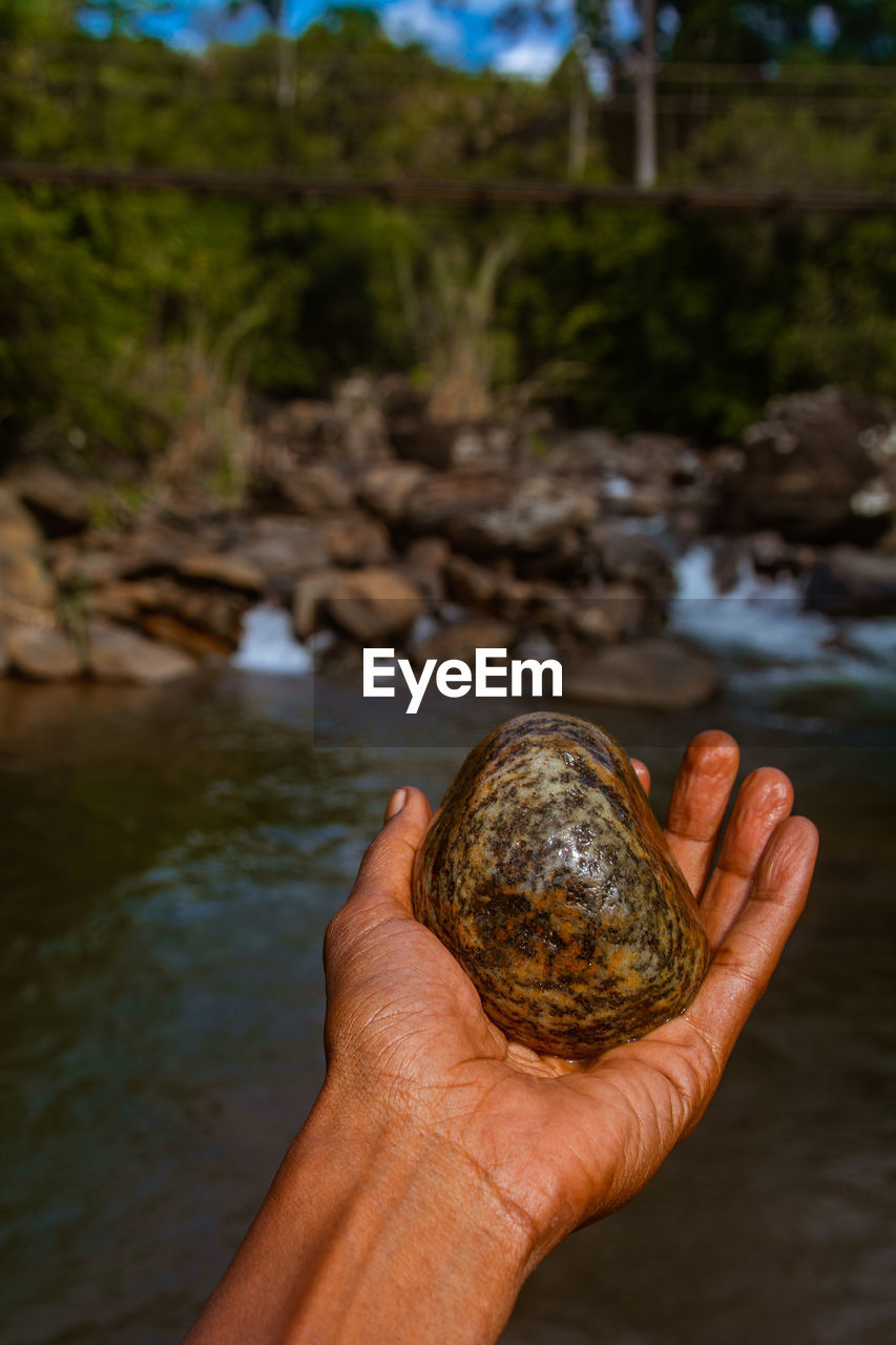 Human hand holding rock against river water and stones