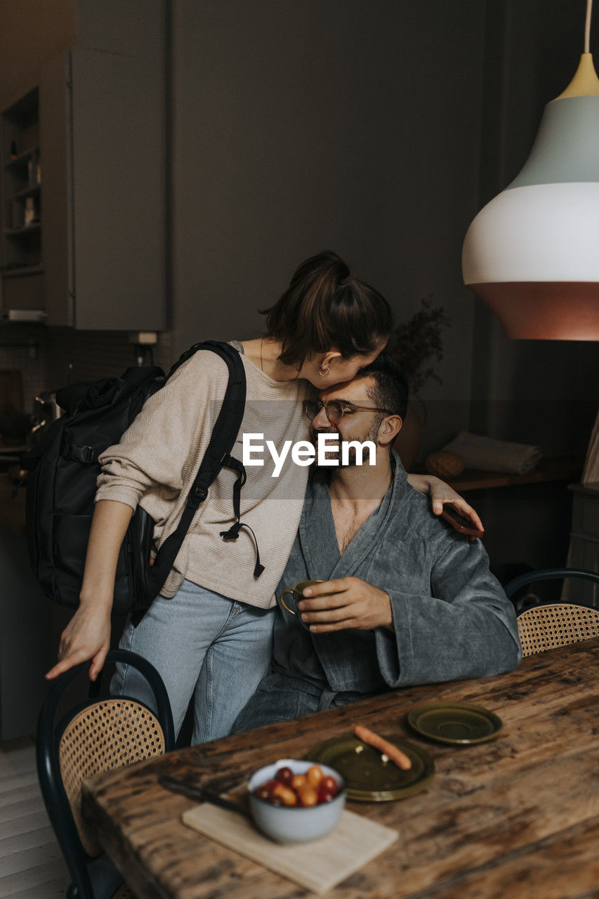 Non-binary person kissing boyfriend sitting at dining table in room