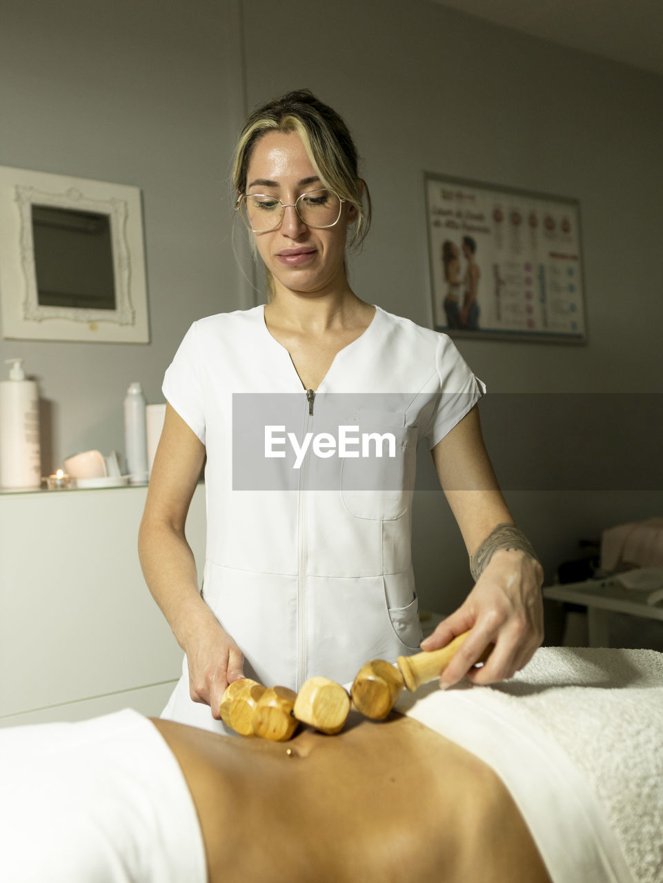 Female masseur with wooden massager doing maderoterapia massage on client belly near towels in light spa salon