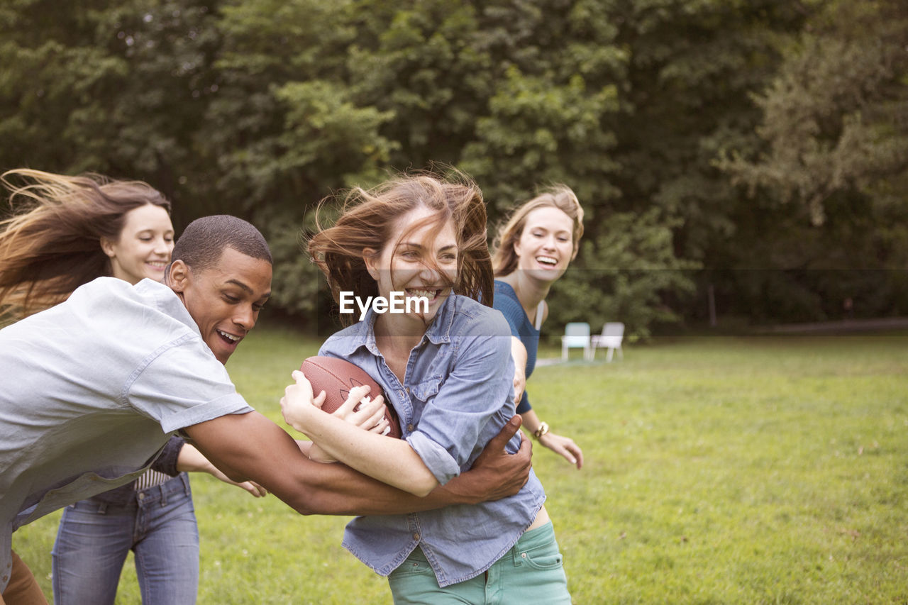 Man pulling woman holding football ball while friends running in background on field