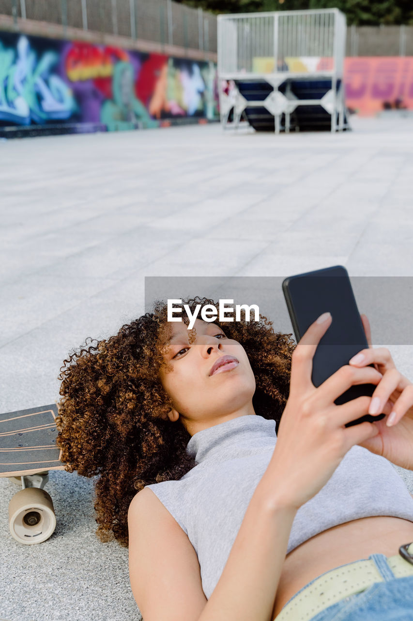 High angle of cheerful black woman with afro hair lying on skateboard on paved ground and using phone to take selfie in town