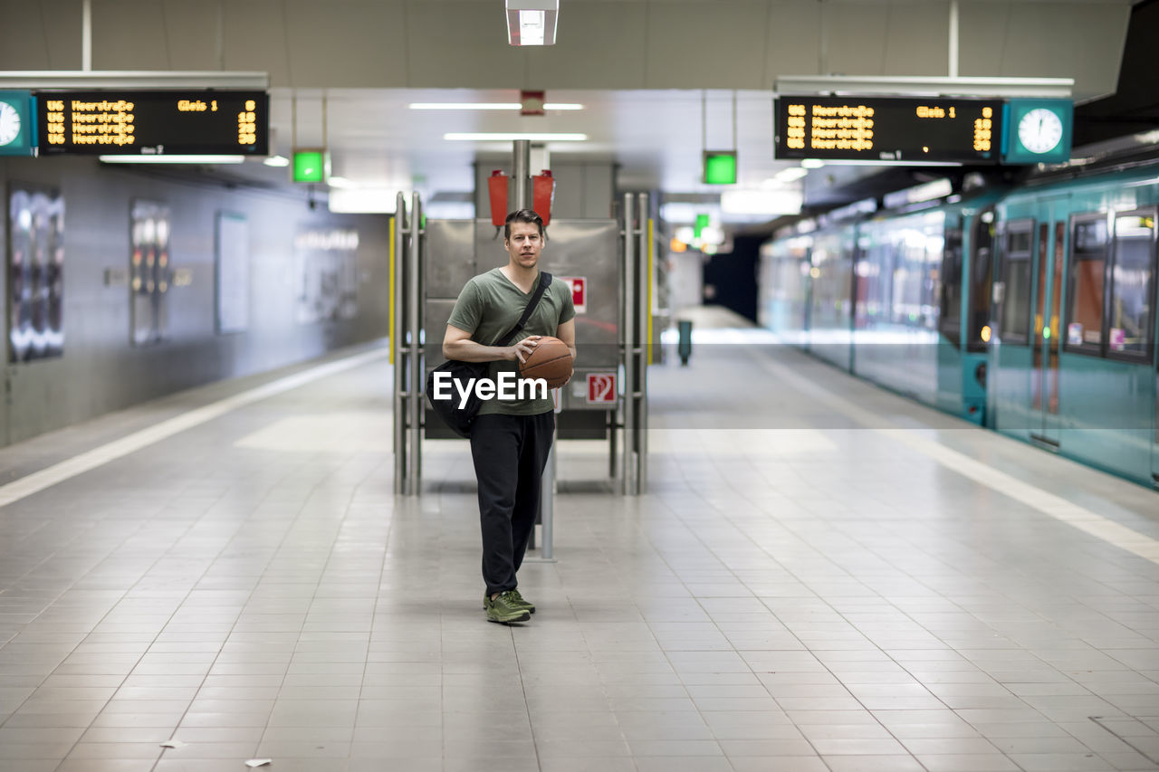 Portrait of young man with basketball walking at railroad station platform