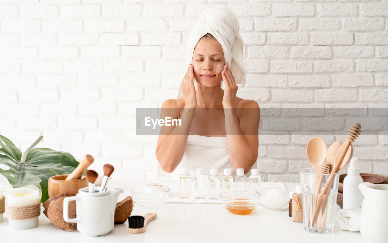 Happy young woman wearing white bathrobes towels on head doing spa procedures applying creme 