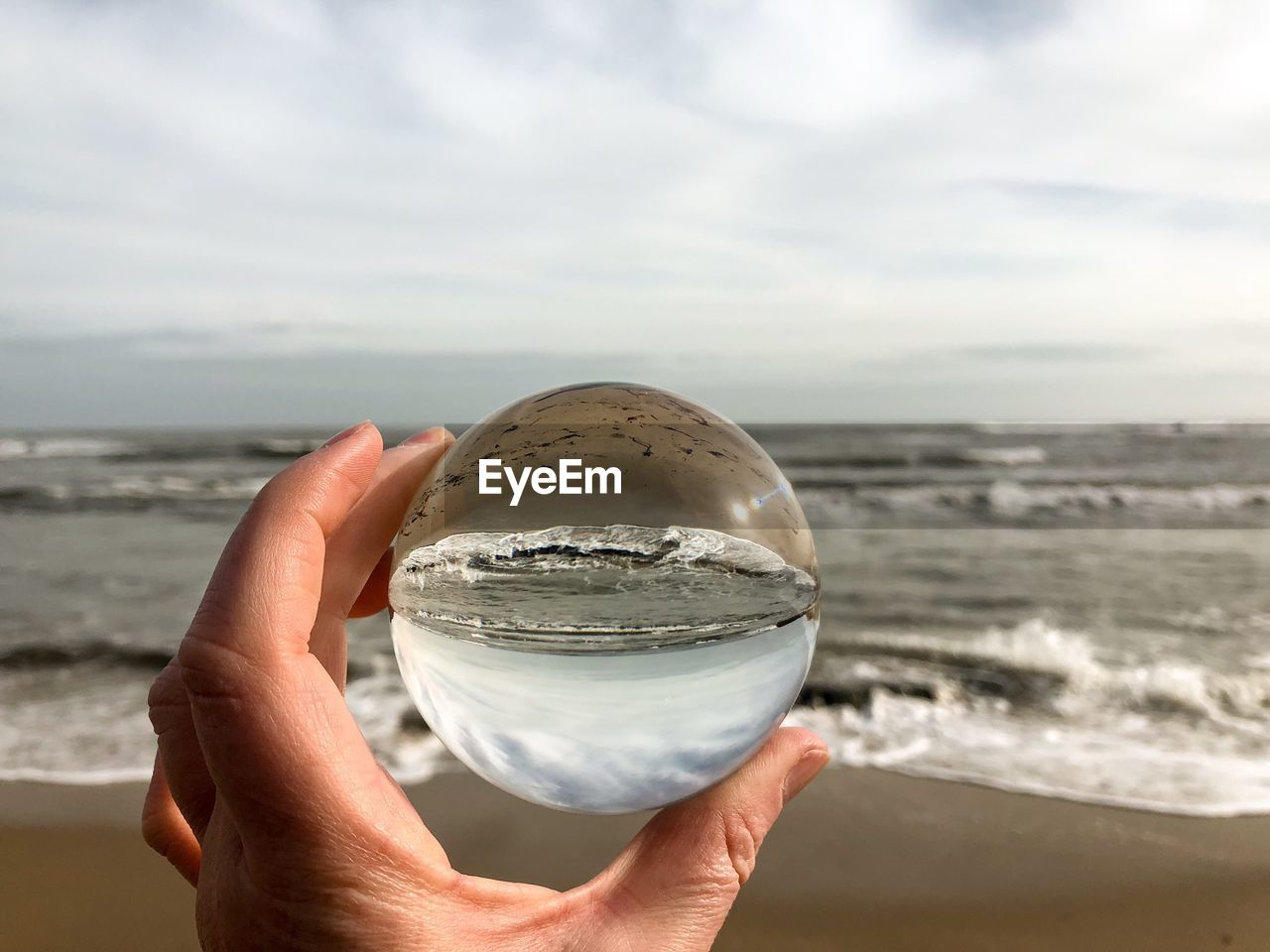 Cropped hand holding crystal ball by sea against cloudy sky