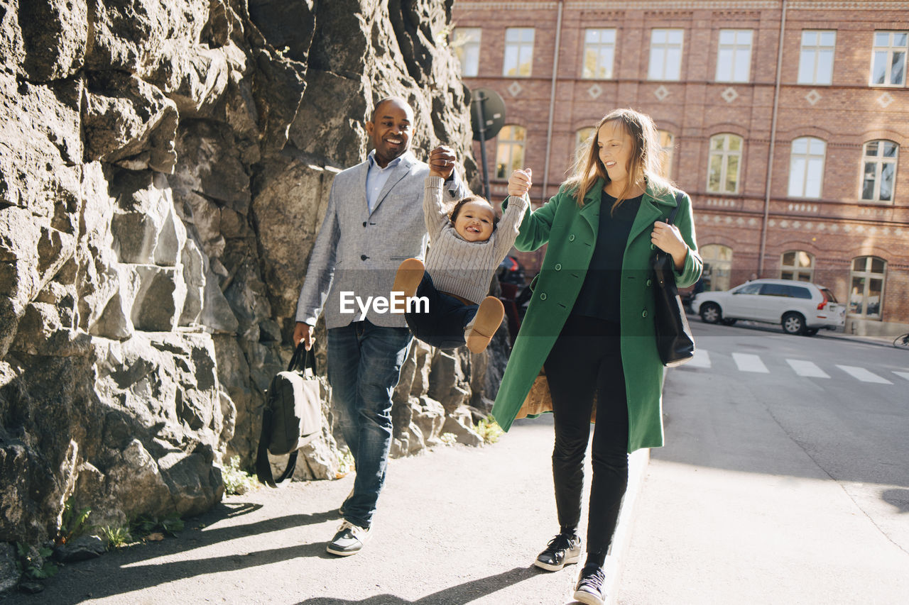 Full length of parents holding daughter in mid-air while walking on sidewalk at city