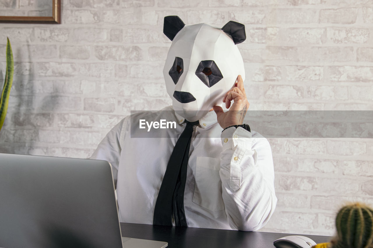 Anonymous tired male entrepreneur in panda bear mask and white shirt working at table with netbook in workspace