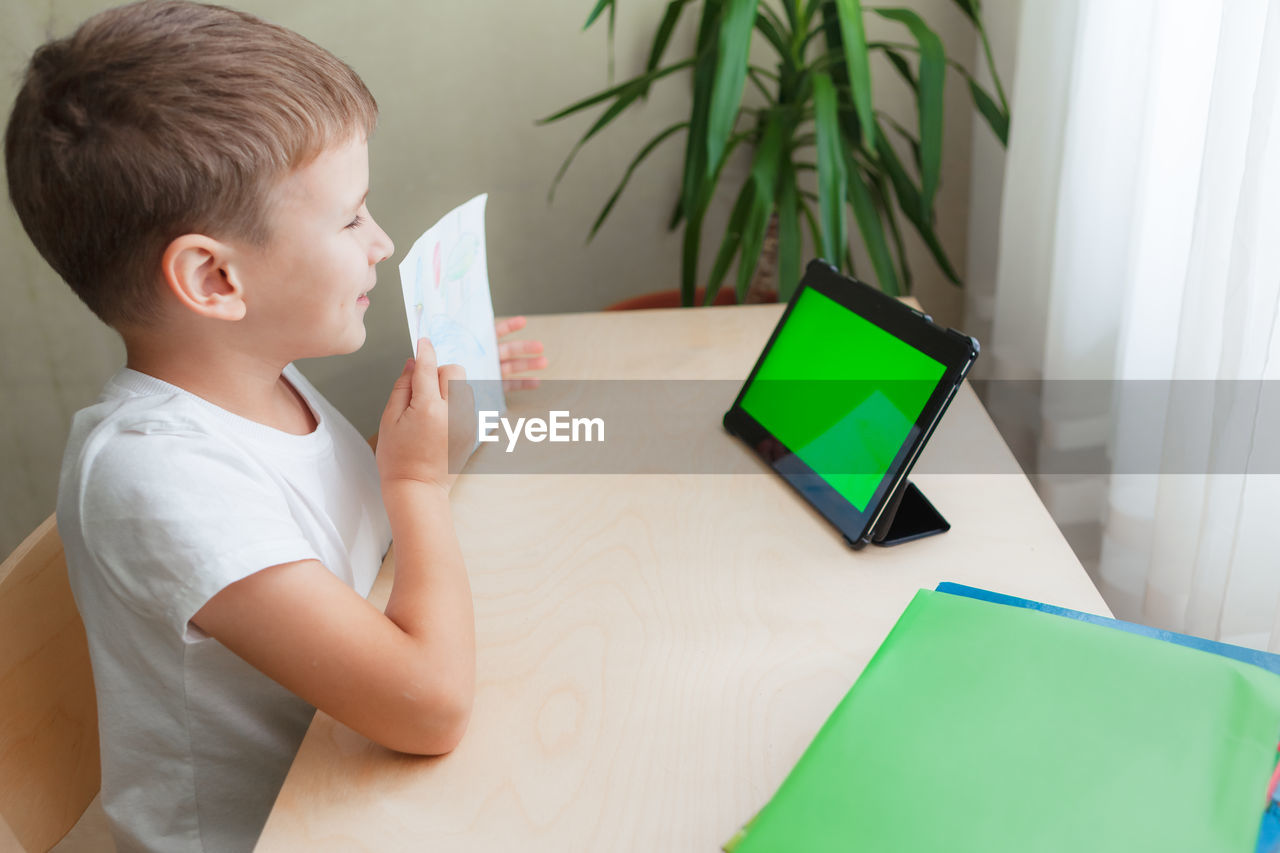 Smiling school boy siting at desk and looks at tablet with green screen.  side view