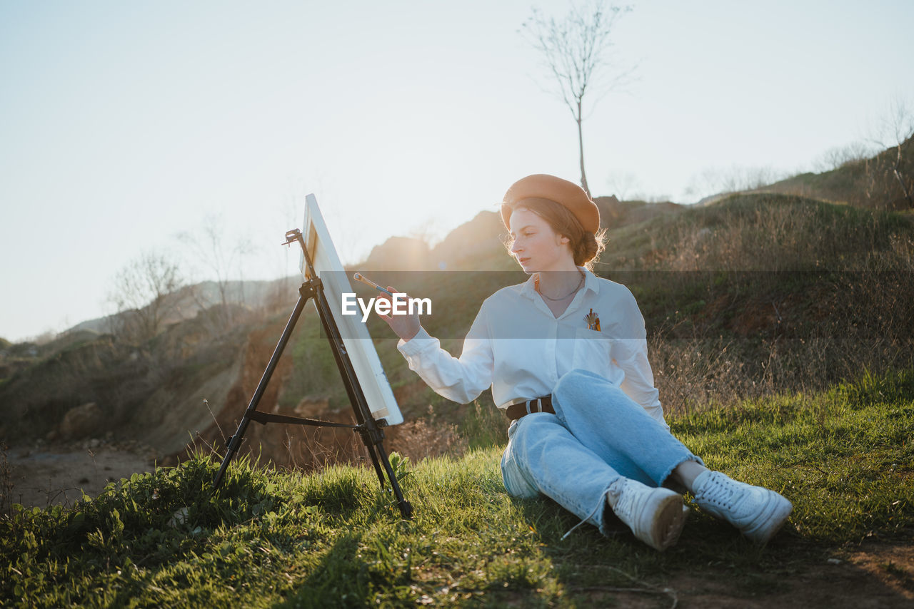 Full body of young woman in beret and stylish clothes sitting on grassy lawn near hills while painting picture with brush on canvas on easel in sunny day under sky