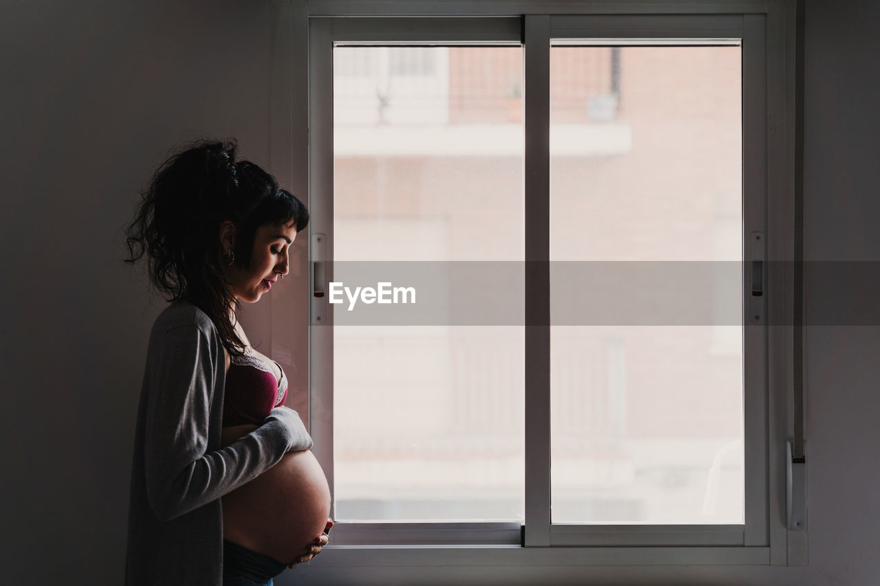 Pregnant woman in bra standing by window at home