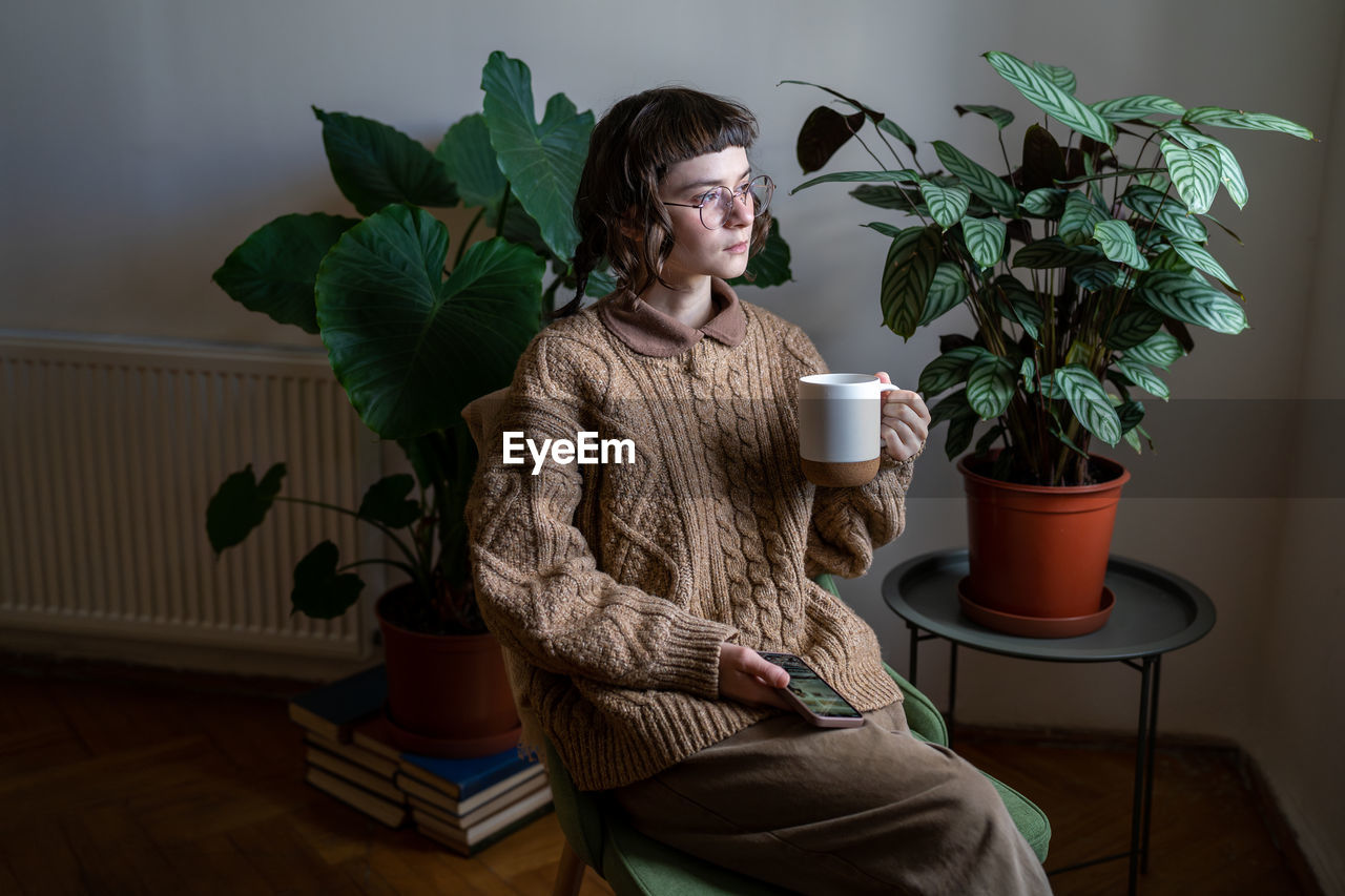 Pensive thoughtful young stylish woman in glasses holding mug looking at window at home with plants.