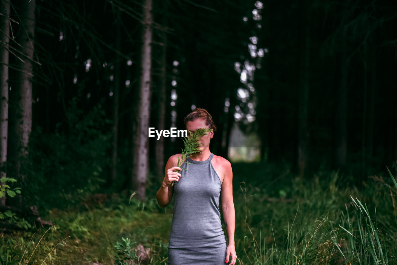 Woman holding leaves over face while standing on grass in forest