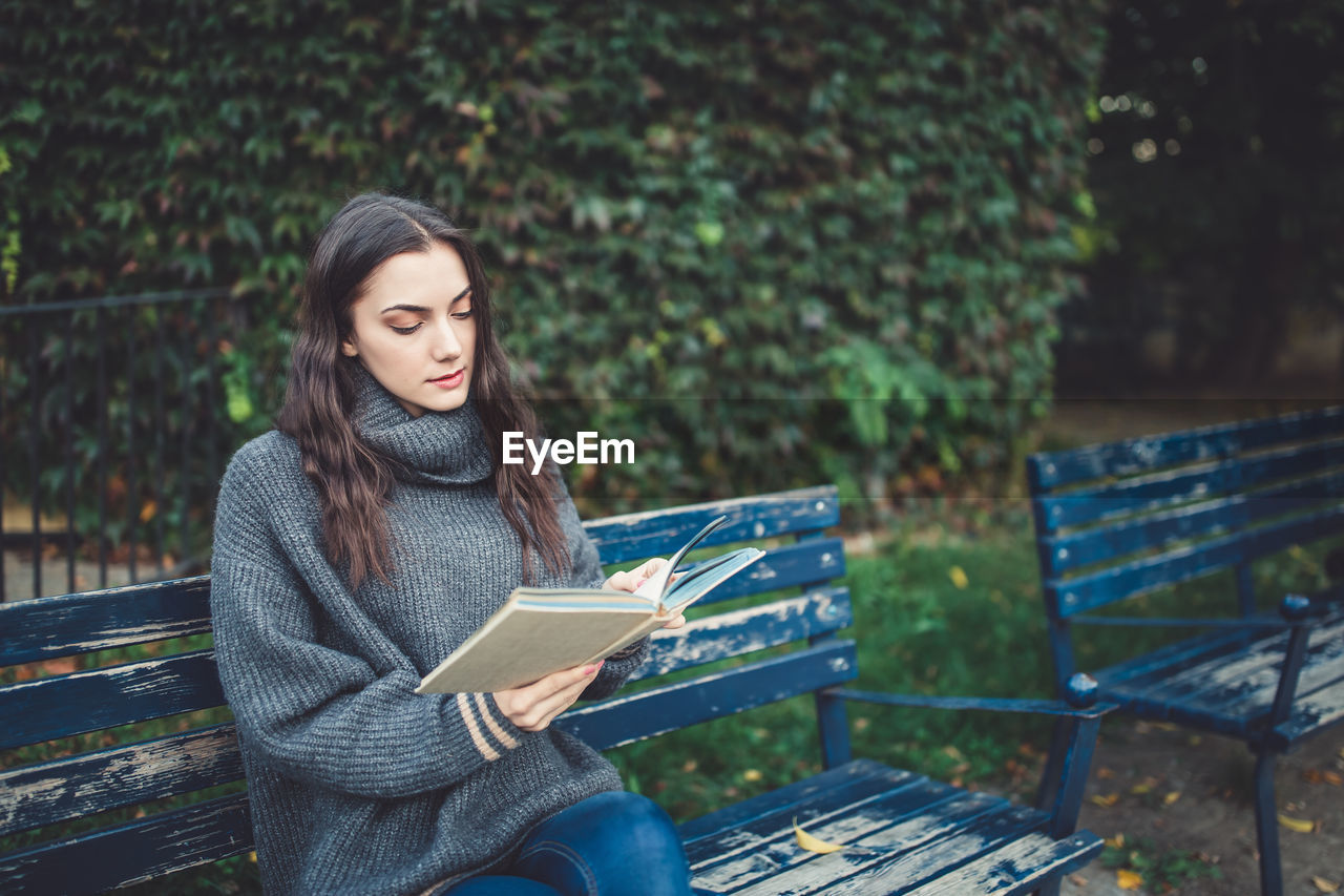 Young woman in a sweater reading a book, sitting on the bench in the park. fall or spring time.