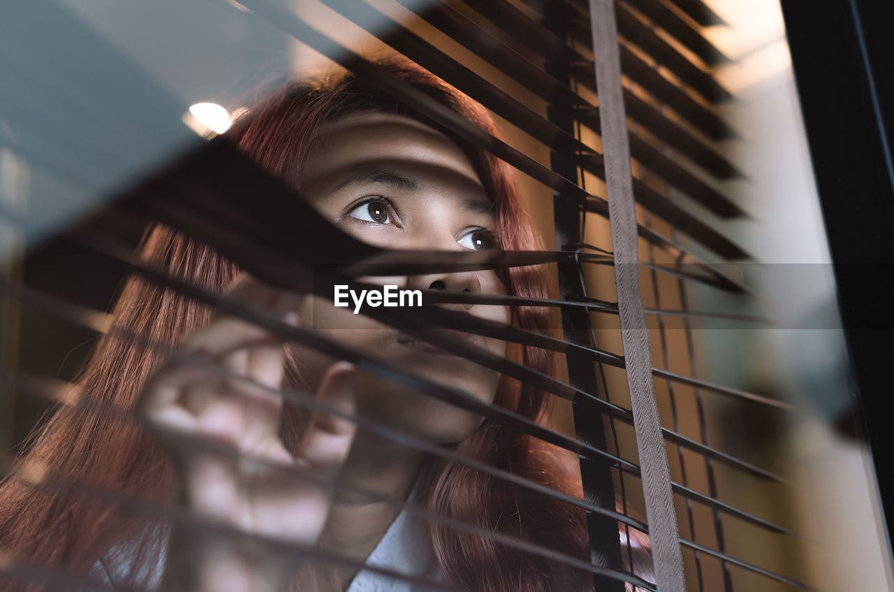 Close-up of woman looking away through blinds and window