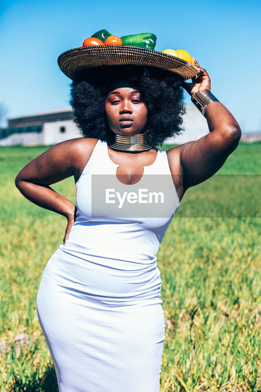 Stylish plus size african woman in white dress with curly hair holding hand on waist and looking at camera while carrying metal plate with bell pepper and tomatoes on head on blurred background of grassy field and blue sky