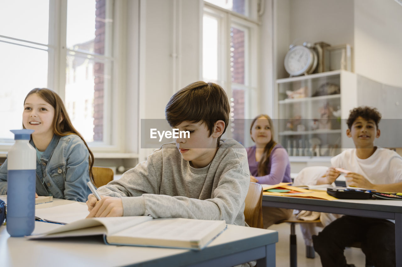 Schoolboy writing in book while sitting at desk with friends in classroom