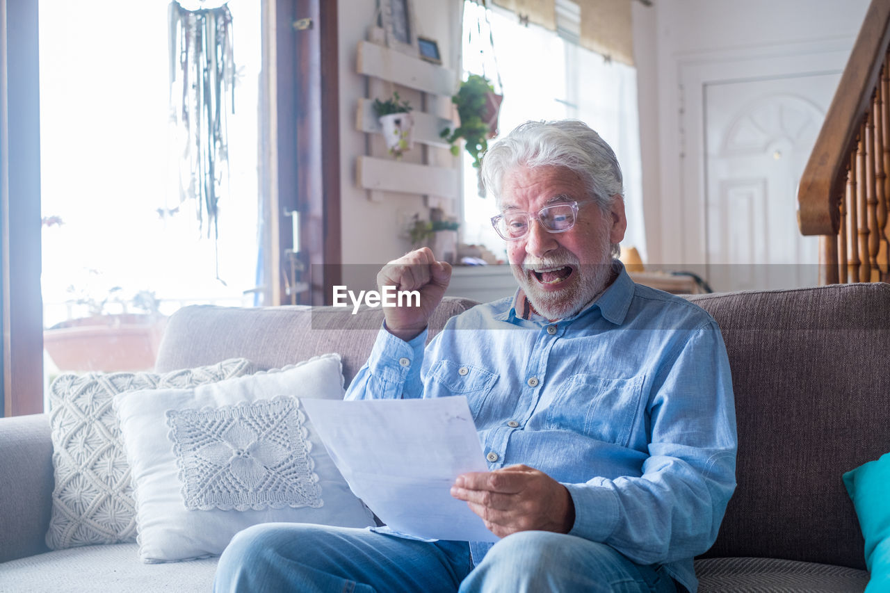 Cheerful senior man holding paper while sitting on sofa at home