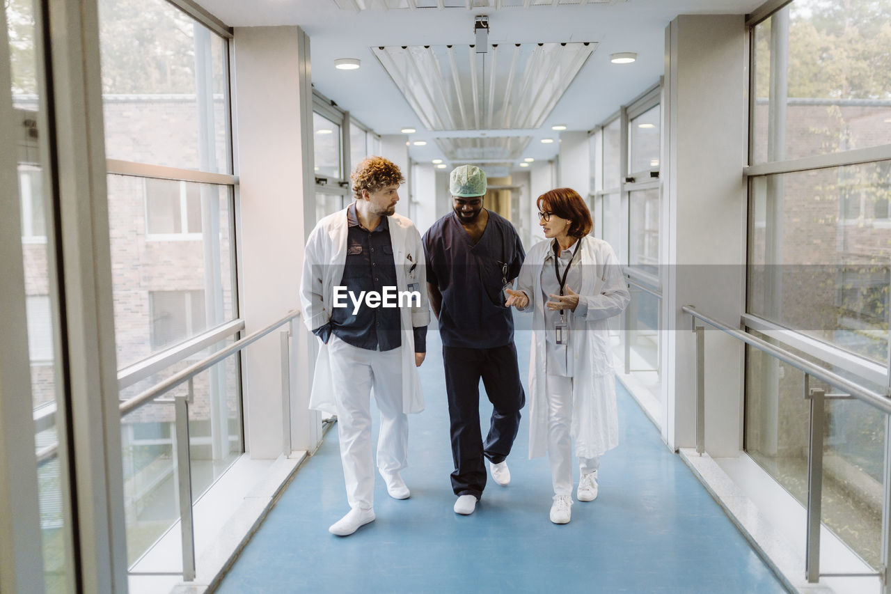 Full length of senior female doctor discussing with colleagues while walking in hospital corridor