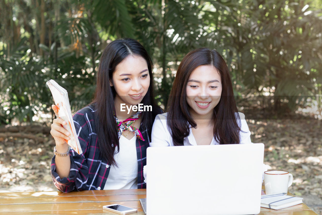 PORTRAIT OF SMILING YOUNG WOMAN USING SMART PHONE WHILE SITTING ON LAPTOP IN PARK