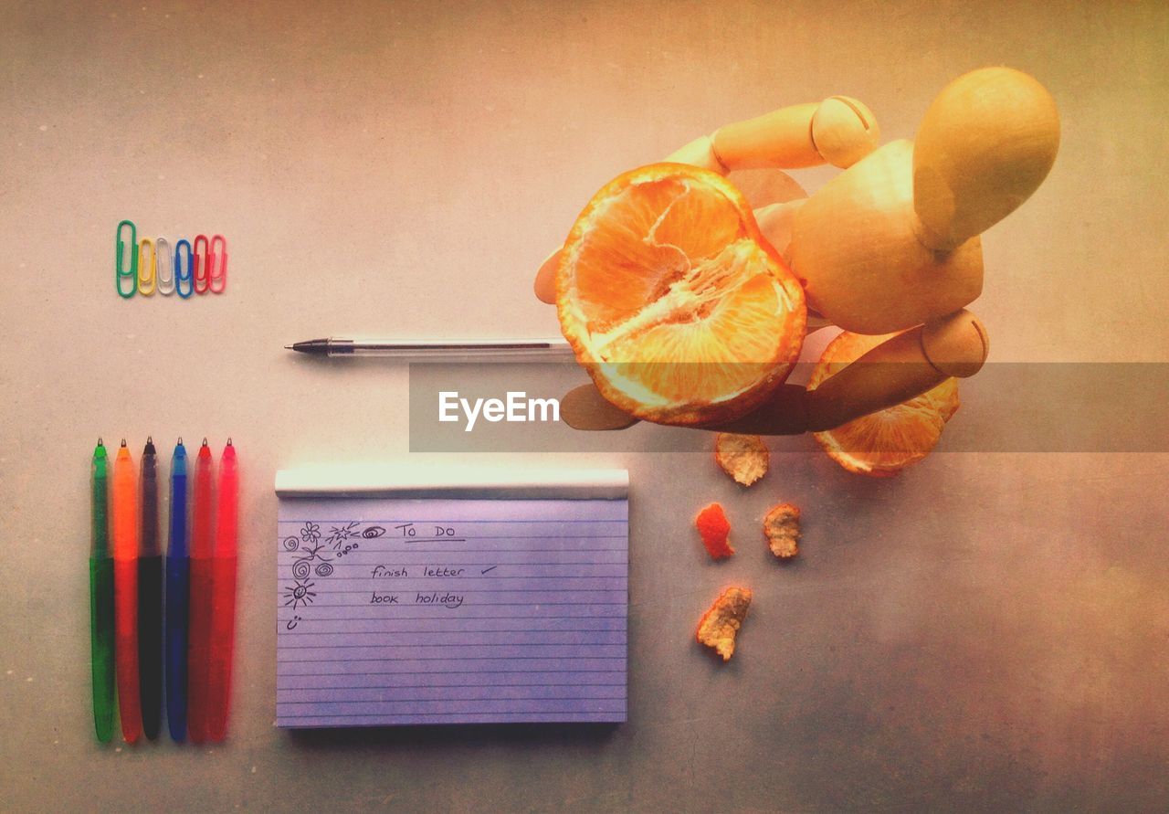 Directly above shot of text on notepad and toy holding orange with colorful pens at desk