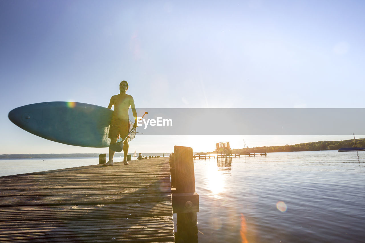 Shirtless man walking with paddleboard on pier over lake against sky