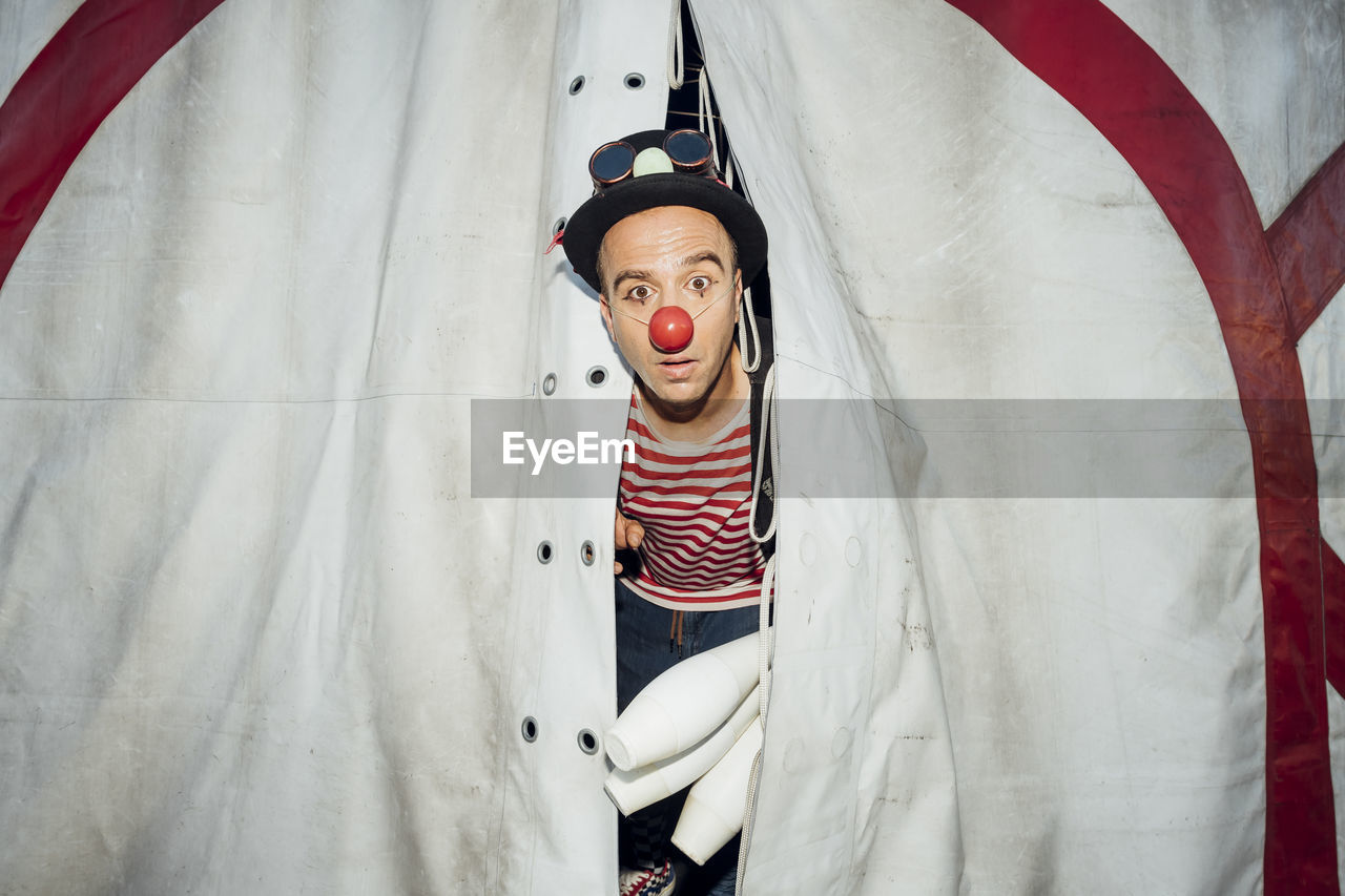Male clown with hat and juggling pin peeking through circus tent entrance