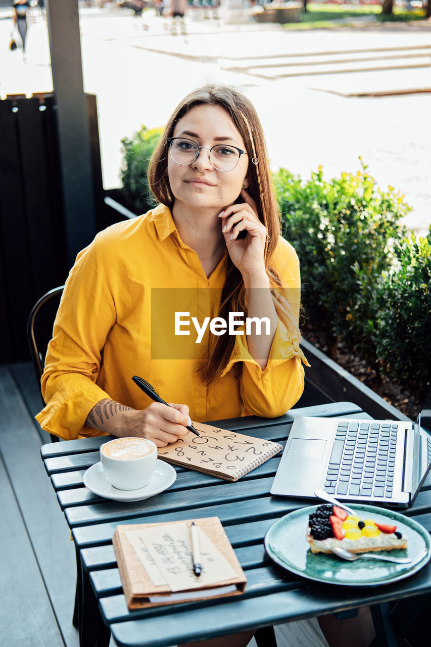 Woman freelancer writing numbers in notepad while working with laptop in street cafe. time