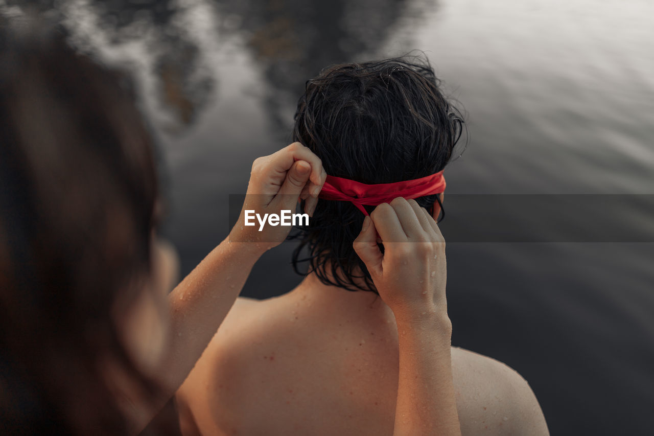 Crop anonymous female tourist tying red blindfold on head of partner against rippled water during trip