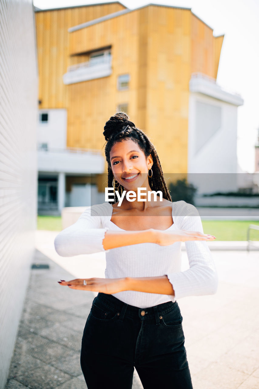 Portrait of young woman gesturing equal sign while standing against building