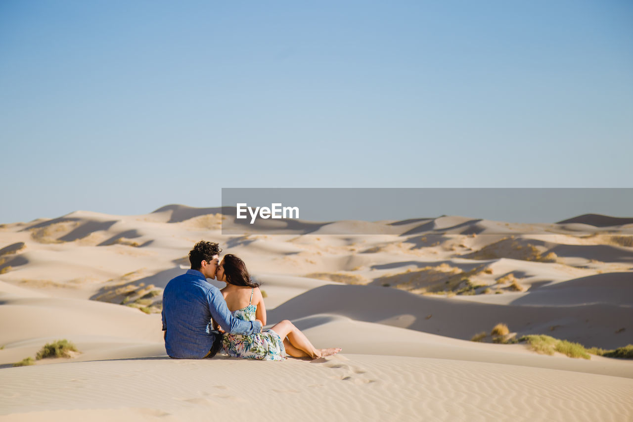 Rear view of couple kissing while sitting on sand at desert against sky